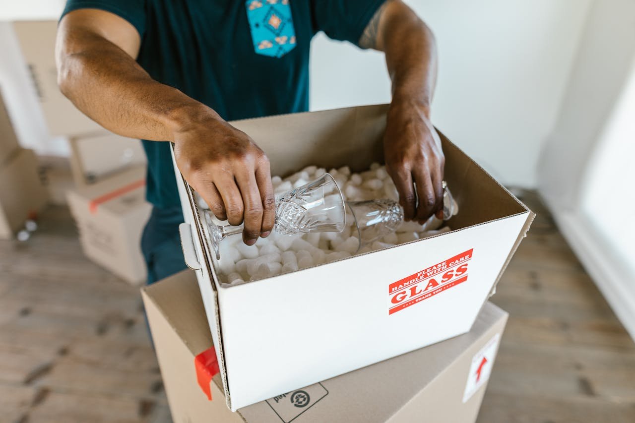 Person packing glassware into boxes with packing peanuts, preparing for relocation.
